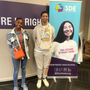 Two students standing in front of a 3DE banner smiling