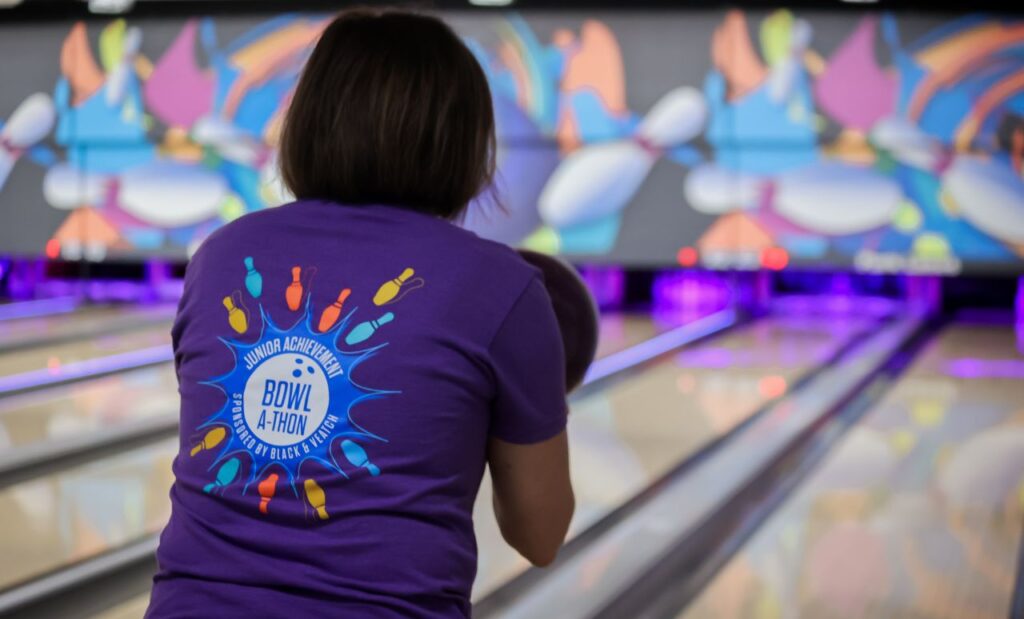 A bowler prepares to throw their ball down the alley, wearing a JA Bowl-A-Thon t-shirt