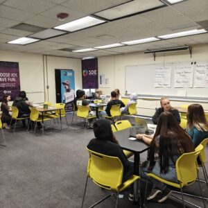 Groups of students in a classroom, sitting at tables talking to a volunteer.