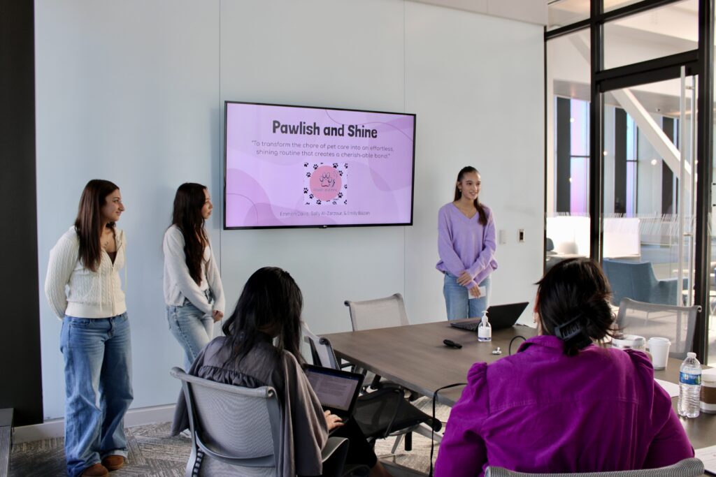 Three students standing by a screen at the front of a conference room giving a presentation.